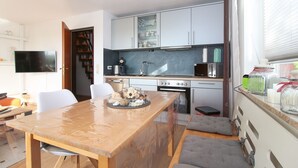 A compact kitchen with modern appliances, a dining area with a wooden table, and a TV mounted on the wall. - Ferienwohnung Seebeere- Obergeschosswohnung in Norddeich für bis zu 4 Personen (Norden)