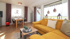 A modern living room with a yellow sofa, a dining area with a wooden table, and large windows with views of the outdoors. - Ferienwohnung Seebeere- Obergeschosswohnung in Norddeich für bis zu 4 Personen (Norden)