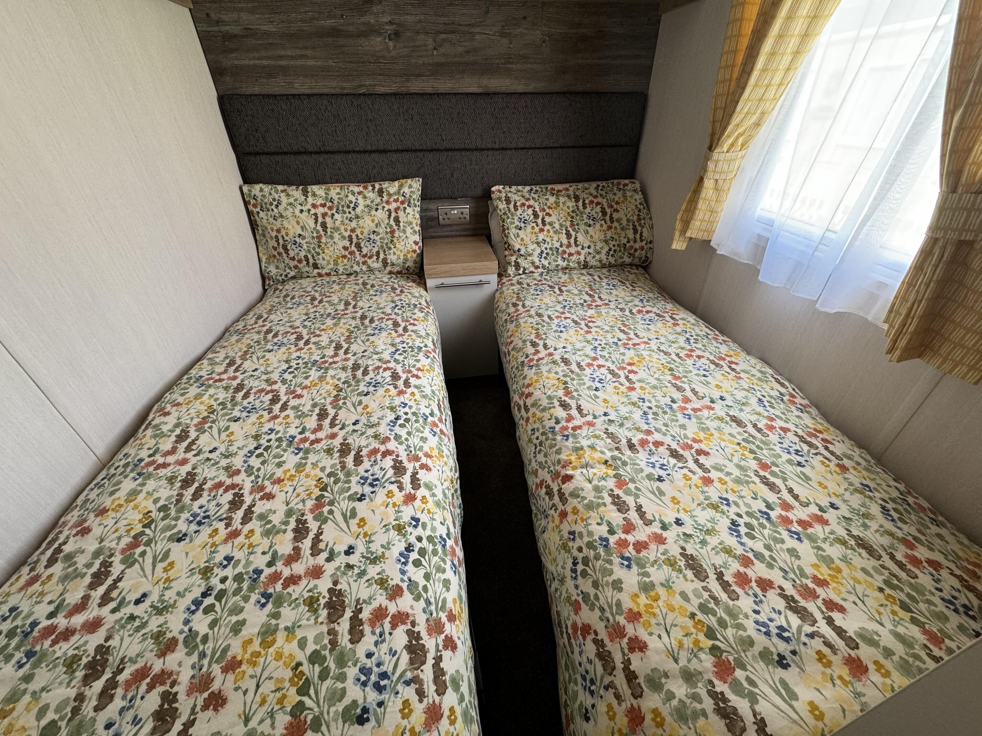 A small, cozy room with two single beds, a wooden headboard, a small table, and a window with sheer curtains.