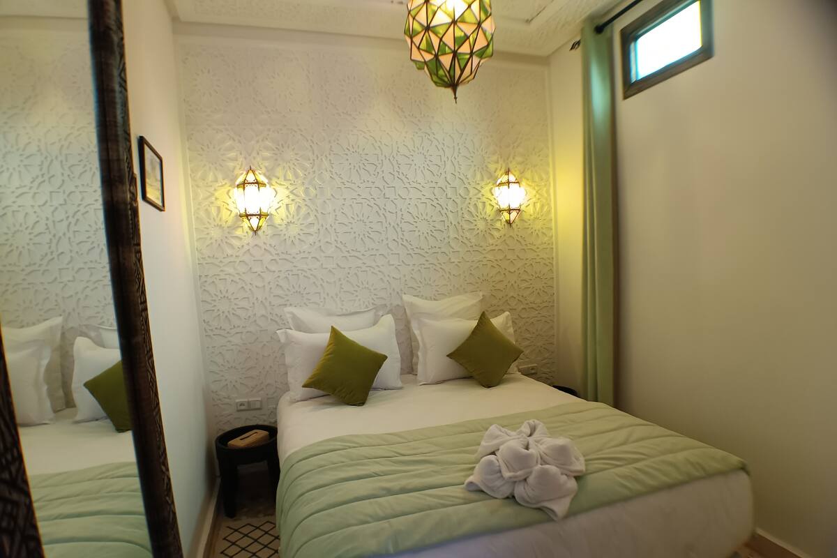 A hotel room with two beds, green bedding, a wall with a geometric pattern, and two wall-mounted lights.