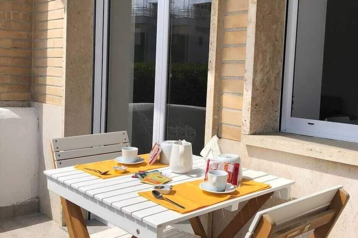 An outdoor patio with a white table and wooden chairs, a yellow table mat, and a window with a view of buildings.