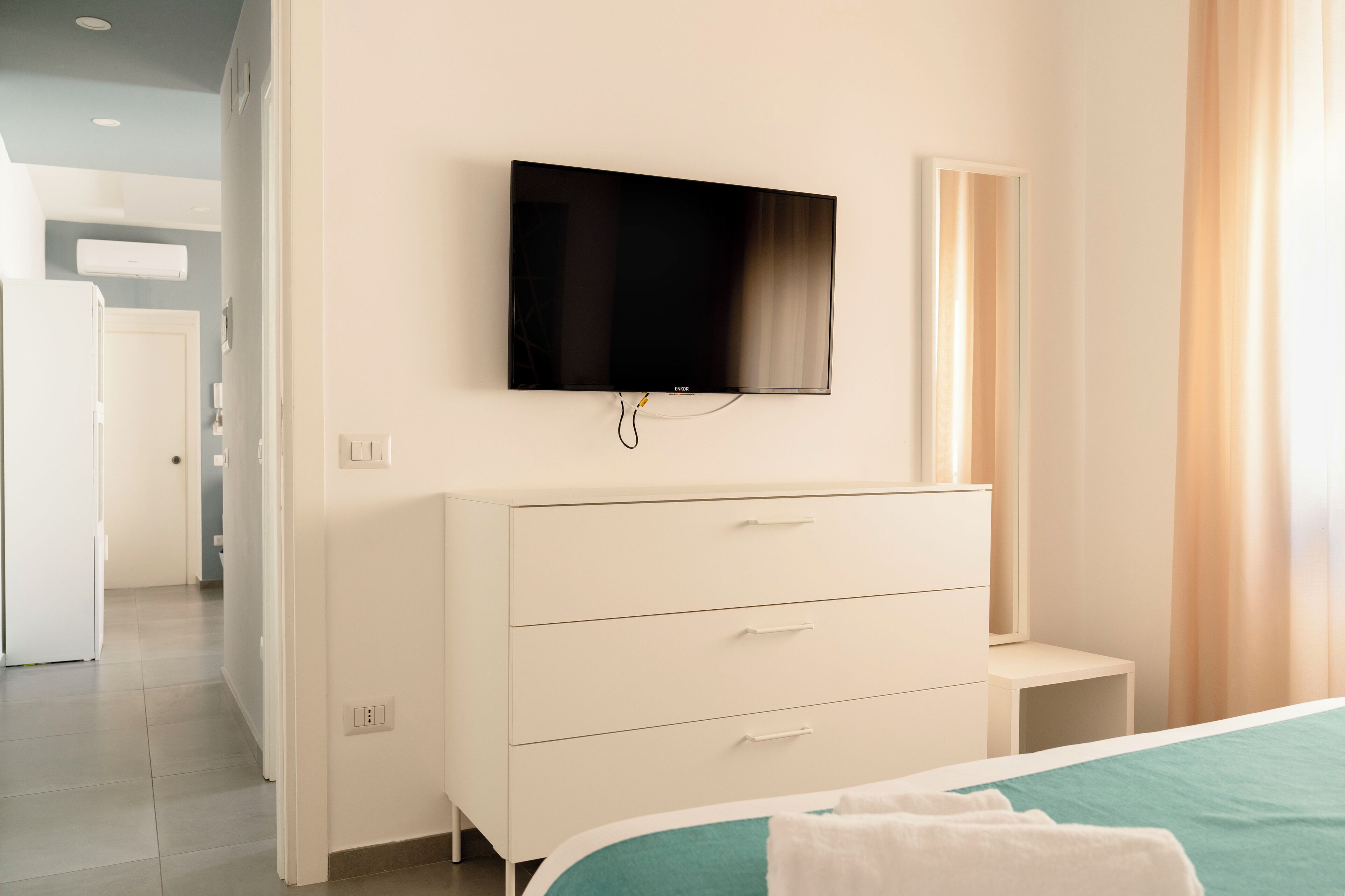 Ein modernes Schlafzimmer mit Flachbildfernseher an der Wand, einer weißen Kommode mit Schubladen und einem Bett mit türkisblau versteckter Bettdecke.