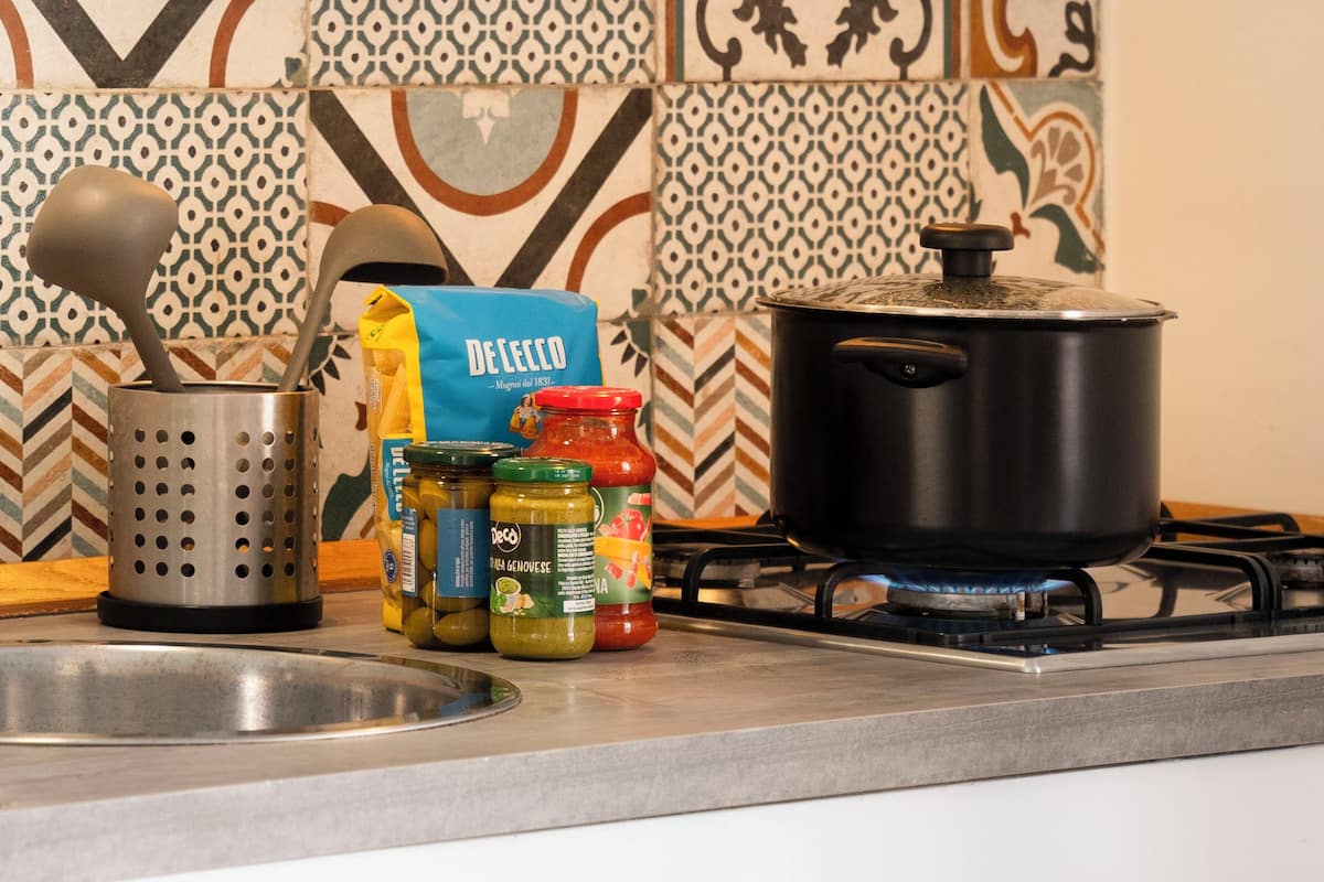 A kitchen with a gas stove, a pot on it, a colander, and jars of condiments.