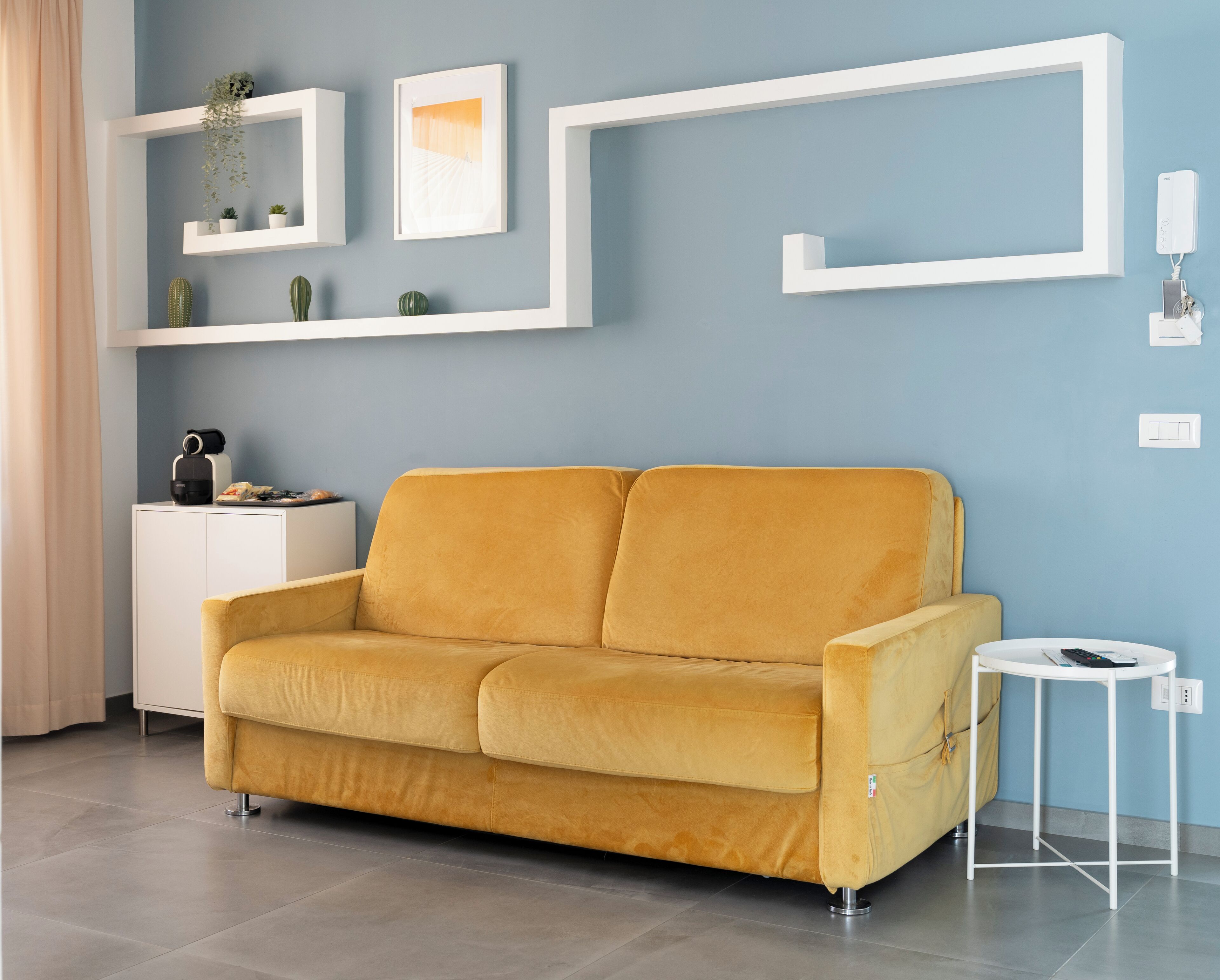 Un canapé de couleur moutarde dans un salon moderne, avec un mur bleu, des étagères blanches et une petite table d’appoint.