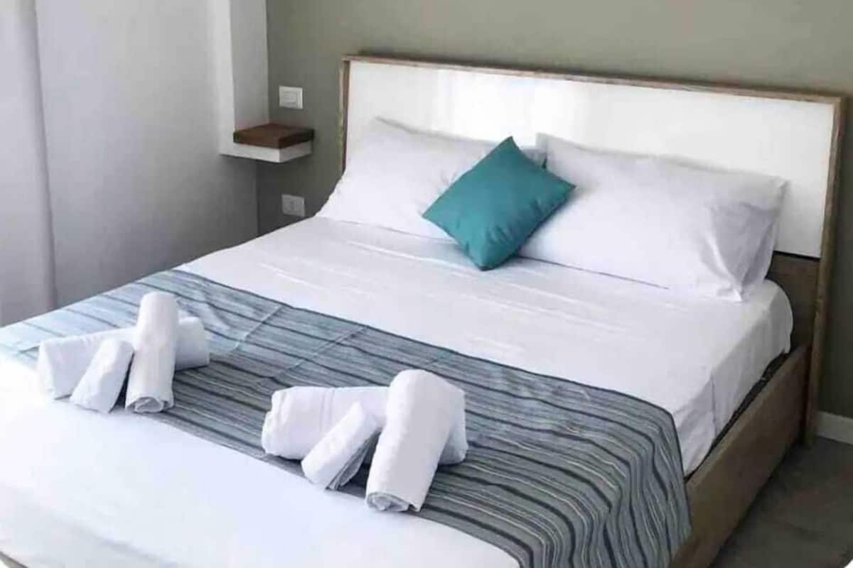 A neatly made bed with white linens and a teal pillow, a wooden headboard, and a small bedside table with a vase.