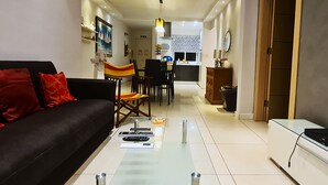 Ein modernes Wohnzimmer mit einer schwarzen Couch, einem Glastisch und einem Essbereich, im Hintergrund eine Küche.