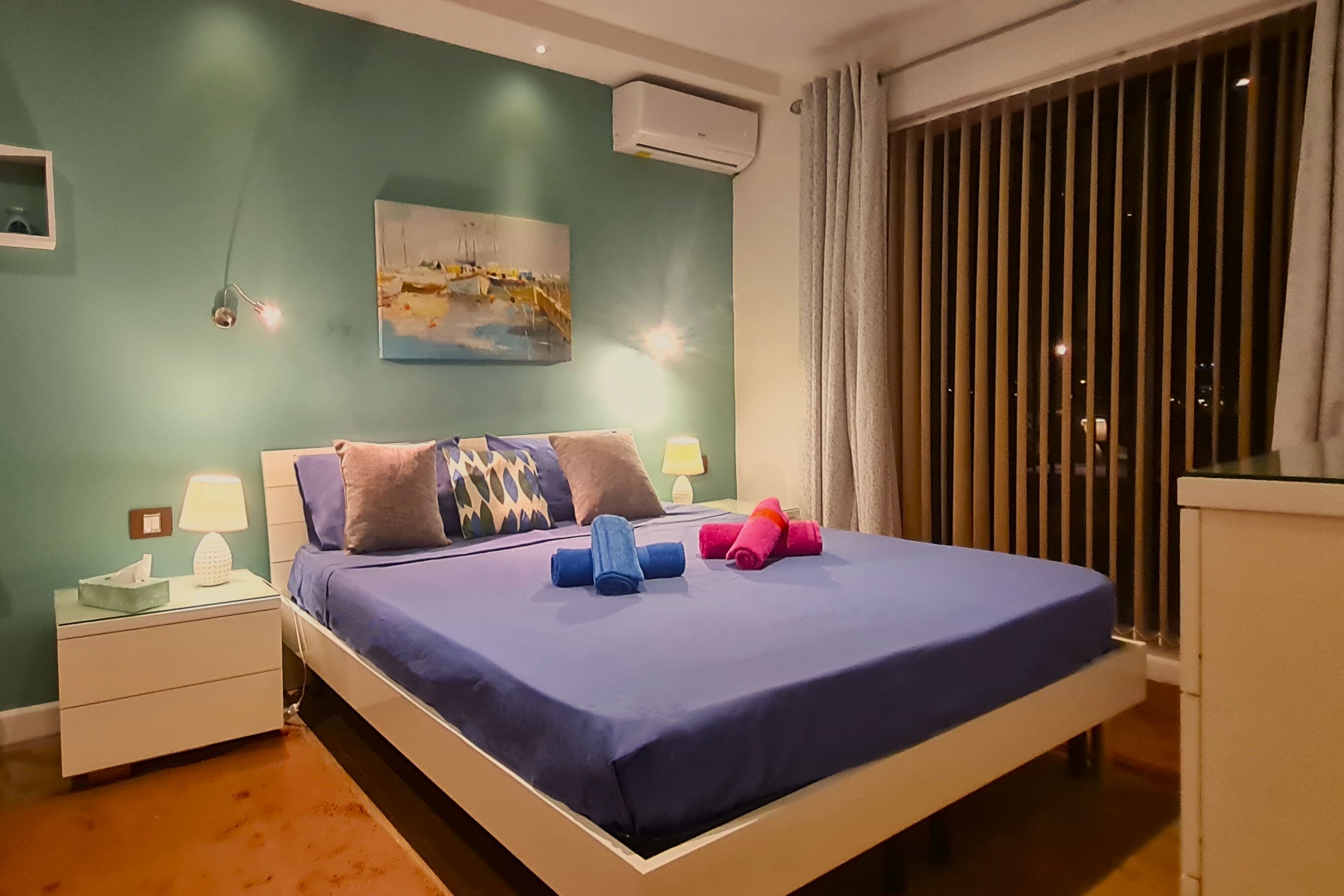 A neatly made bed with blue bedding, a painting on the wall, and a nightstand with a lamp.