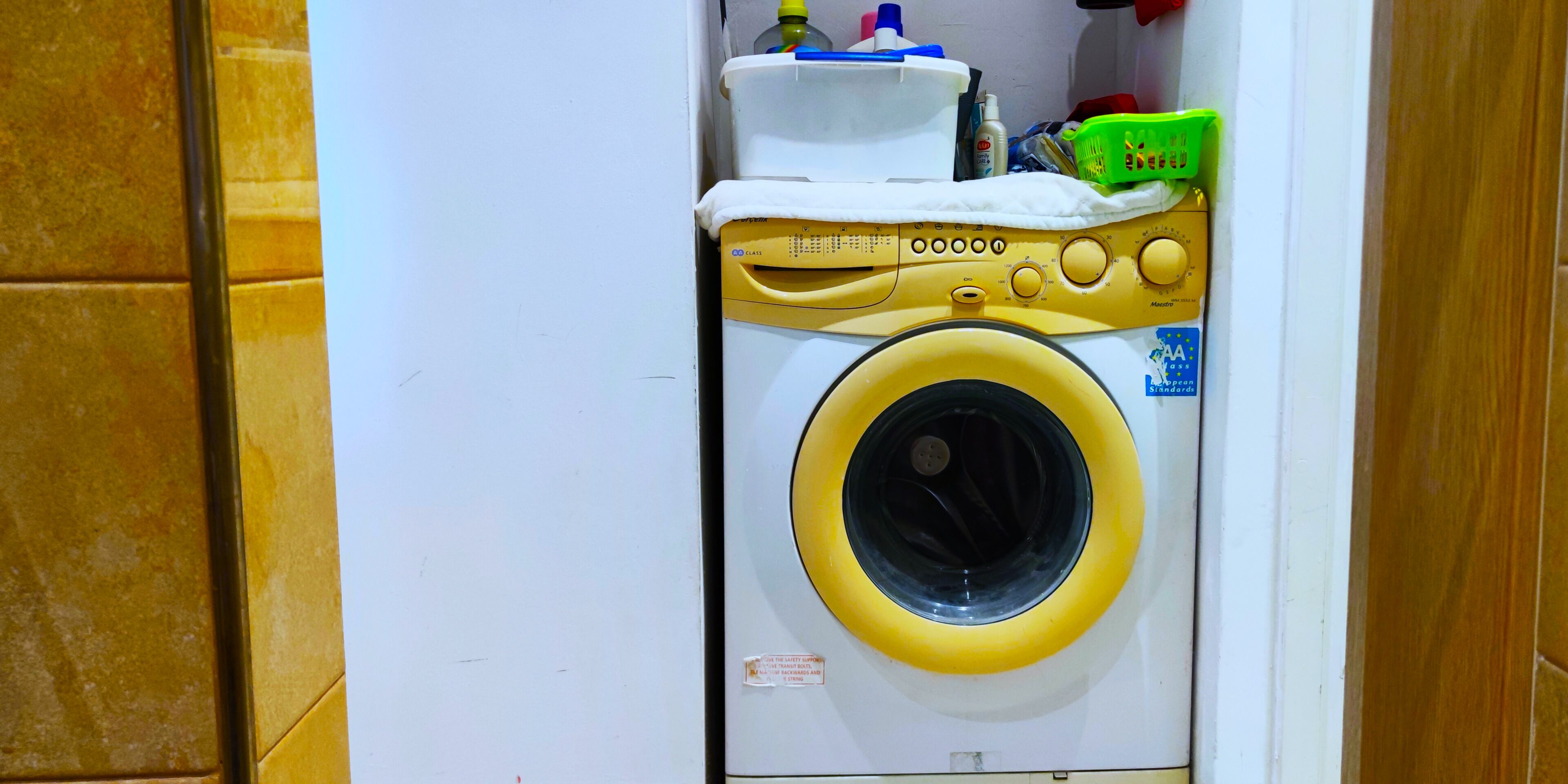 A compact, wall-mounted washing machine with a control panel, a detergent bottle, and a green basket.