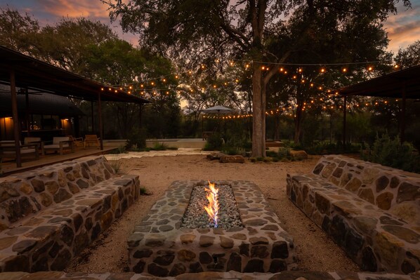 An outdoor seating area with stone benches and a central fire feature, surrounded by trees and string lights.