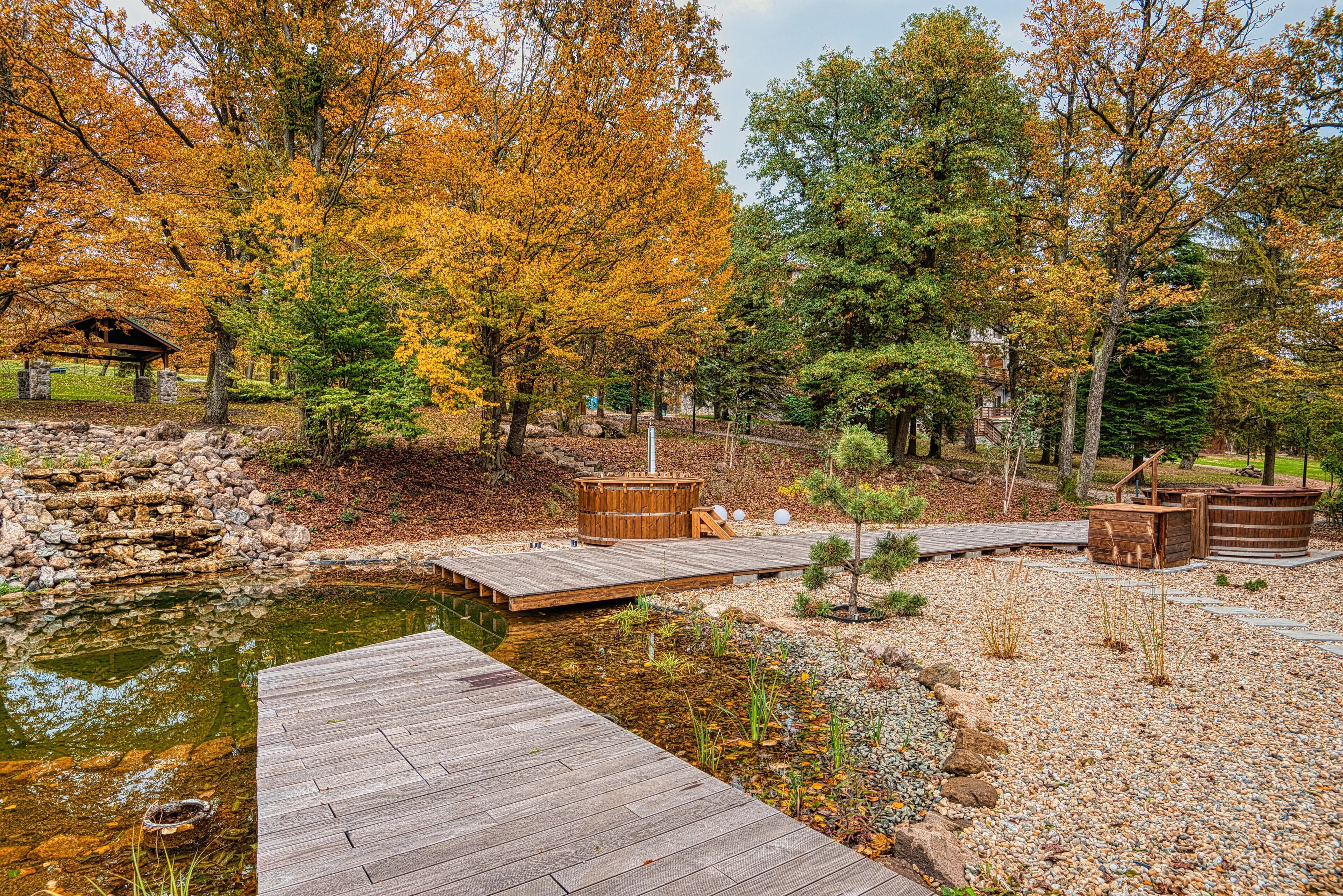 A wooden deck leading to a pond with a wooden gazebo and a wooden hot tub.