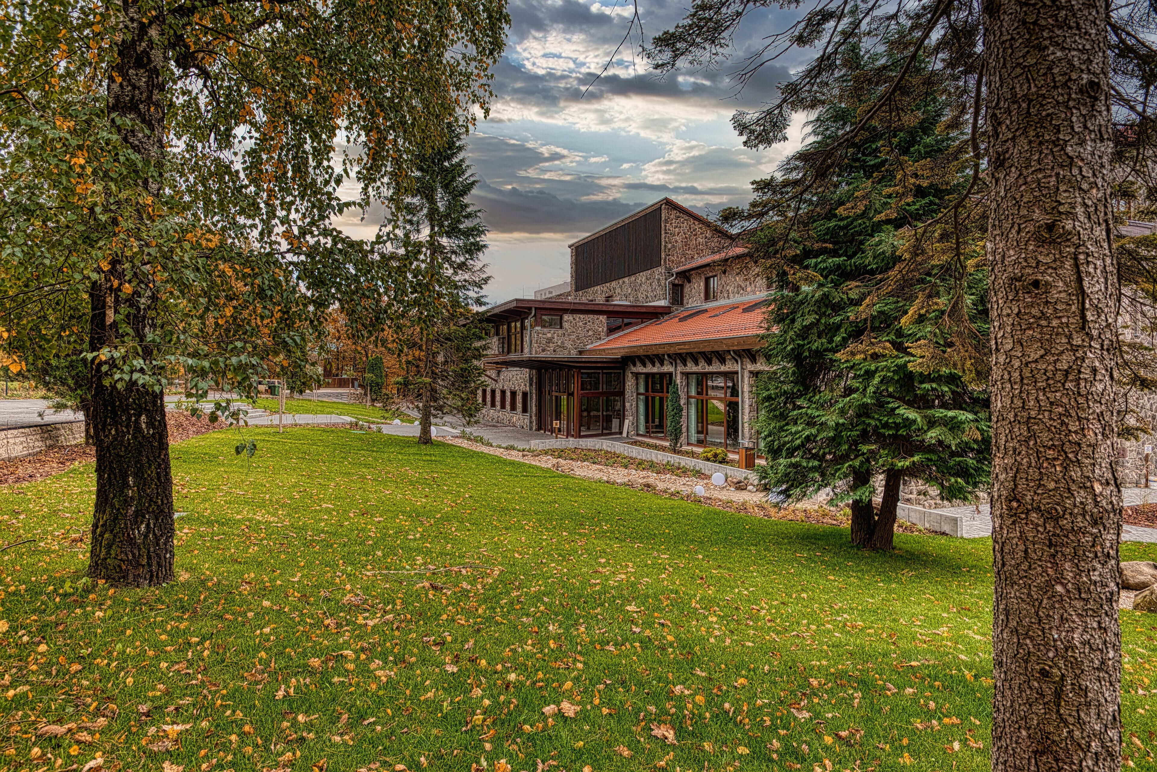 A modern house with a stone facade, large windows, and a red roof, surrounded by trees and a grassy lawn.