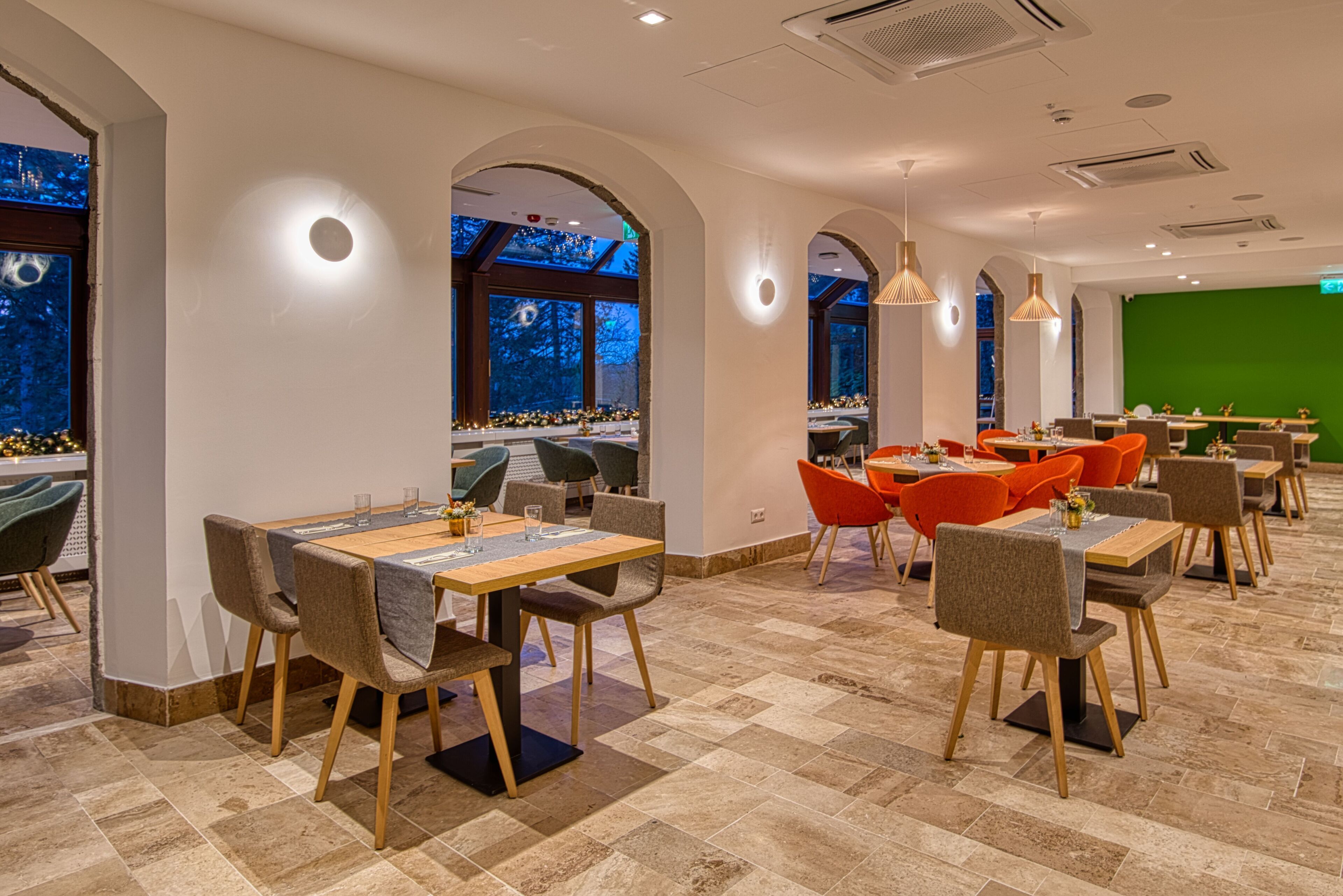 A modern restaurant with a high ceiling, arched windows, and a green screen in the background. Tables are arranged in a U-shape, and the floor is tiled.