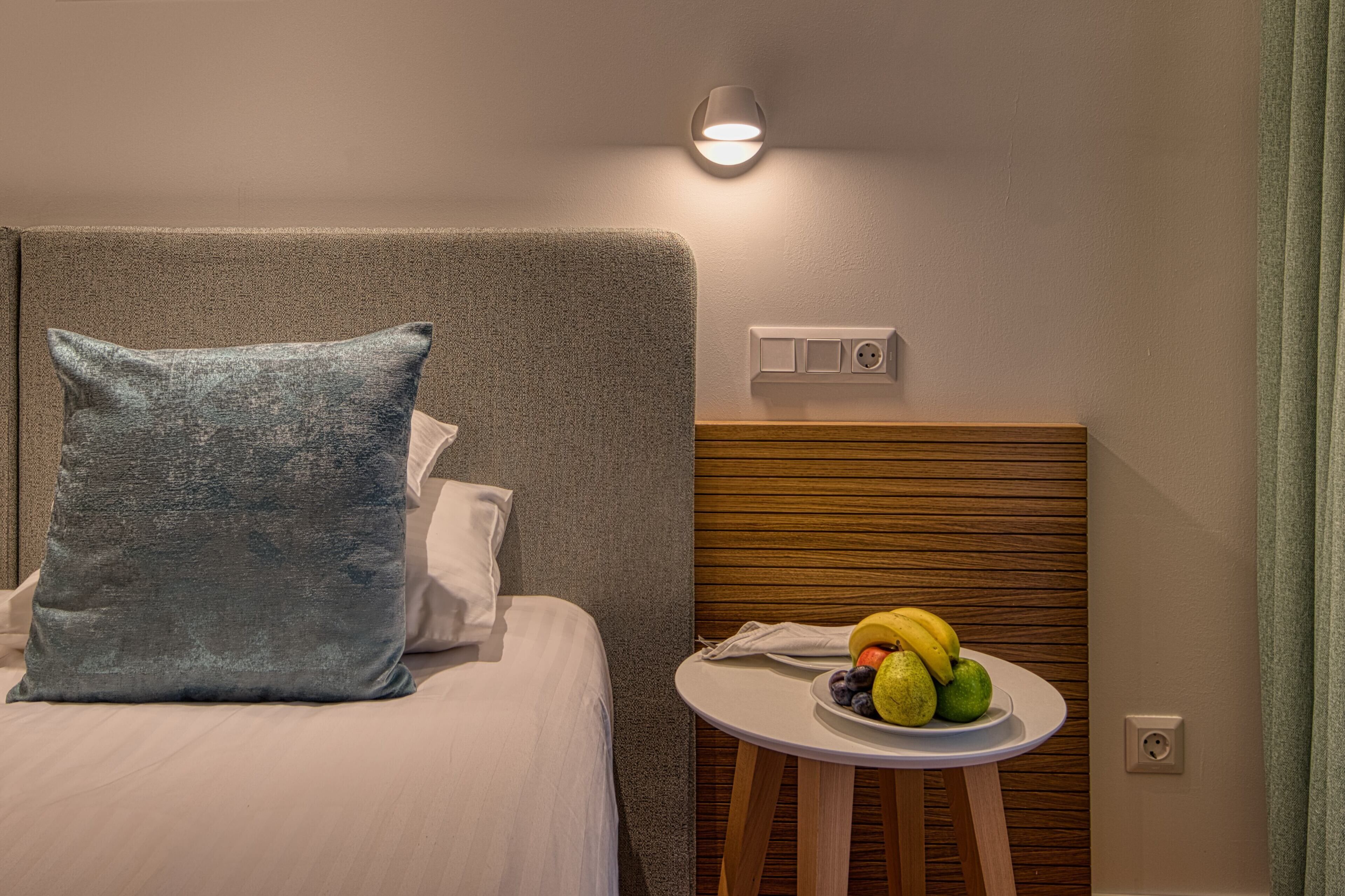 Habitación de hotel con cama, mesita de noche con frutas, luz de pared y toma de corriente.