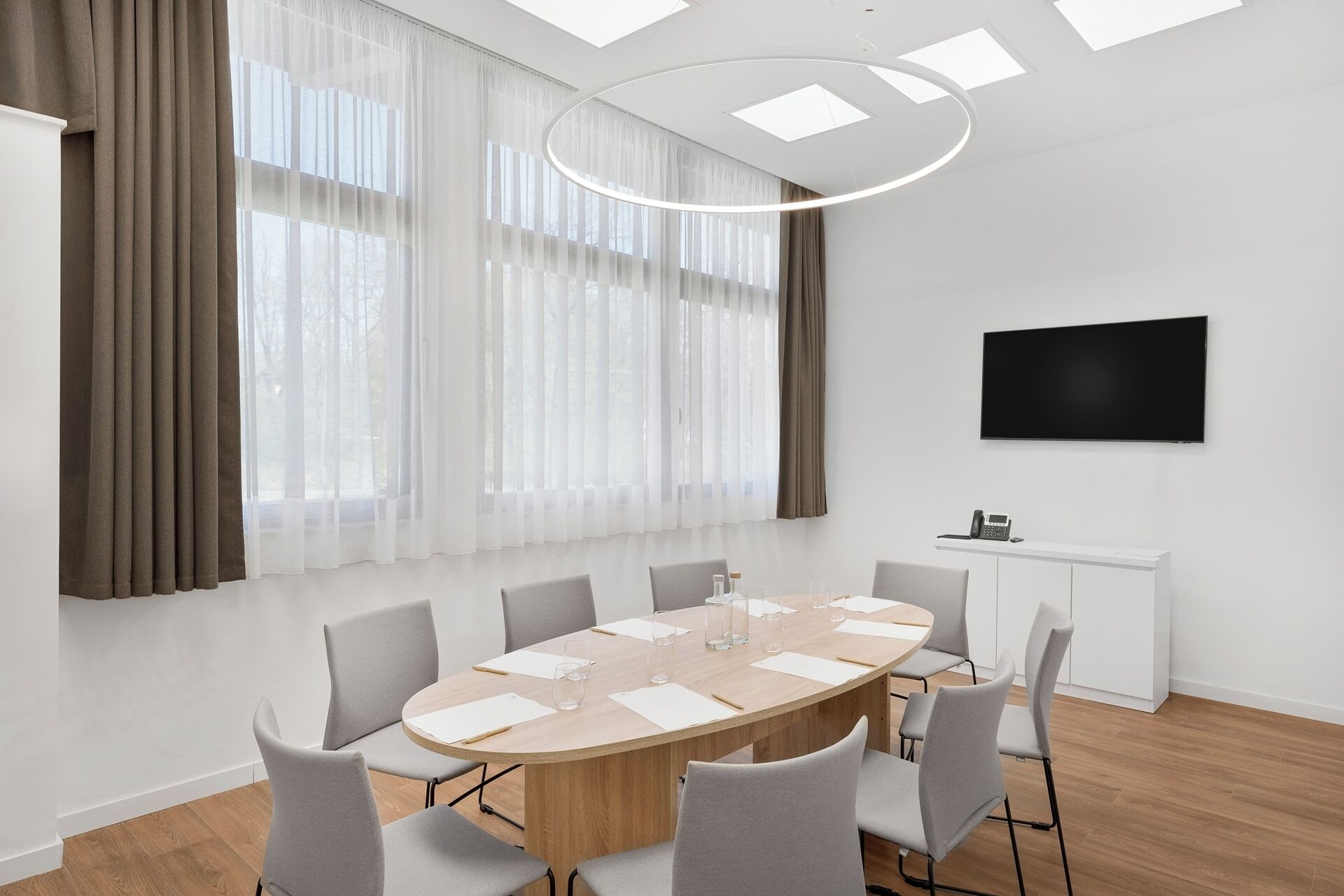 Une salle de conférence équipée d’une table ronde, de chaises, d’un téléviseur fixé au mur et de grandes fenêtres avec des rideaux.