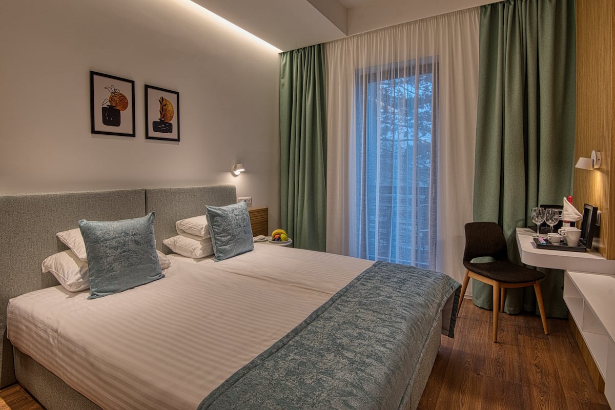 Ein Hotelzimmer mit einem großen Bett, einem Schreibtisch mit Stuhl, einem Fenster mit Vorhängen und zwei gerahmten Bildern an der Wand.