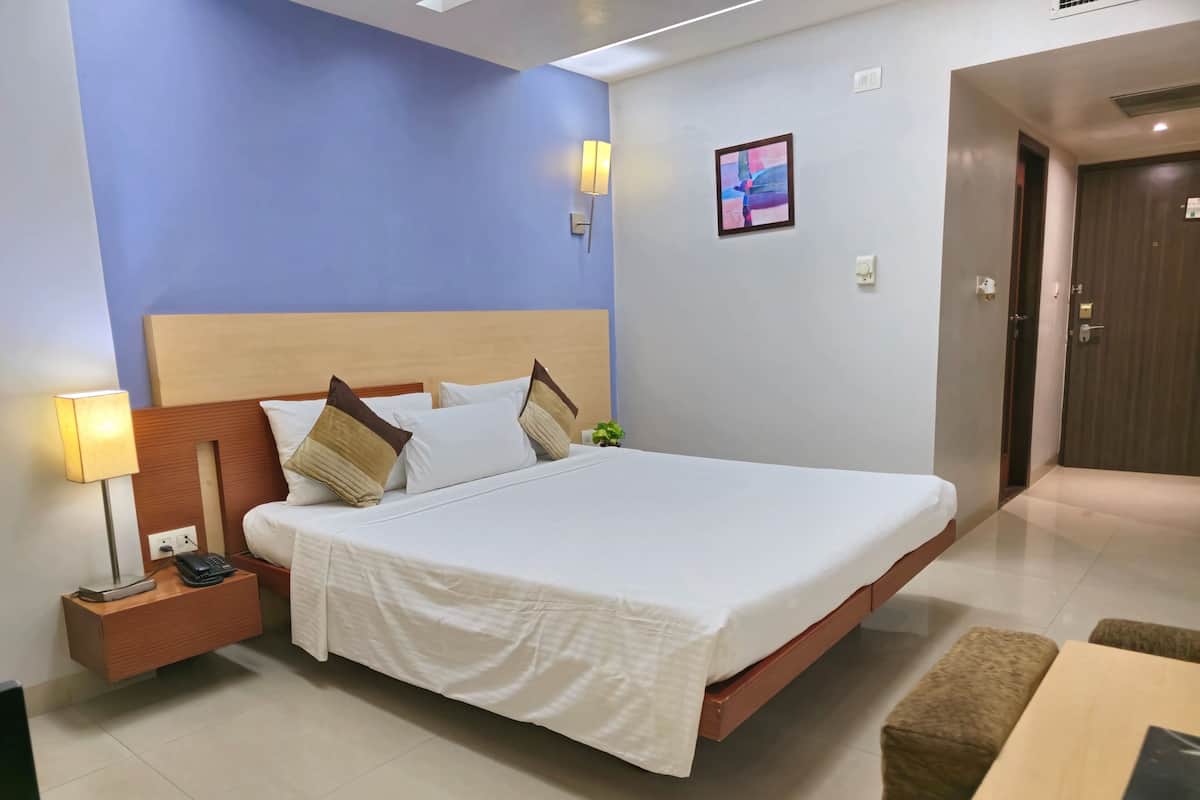 A hotel room with a large bed, bedside table with a lamp, a small plant, and a door to the right.