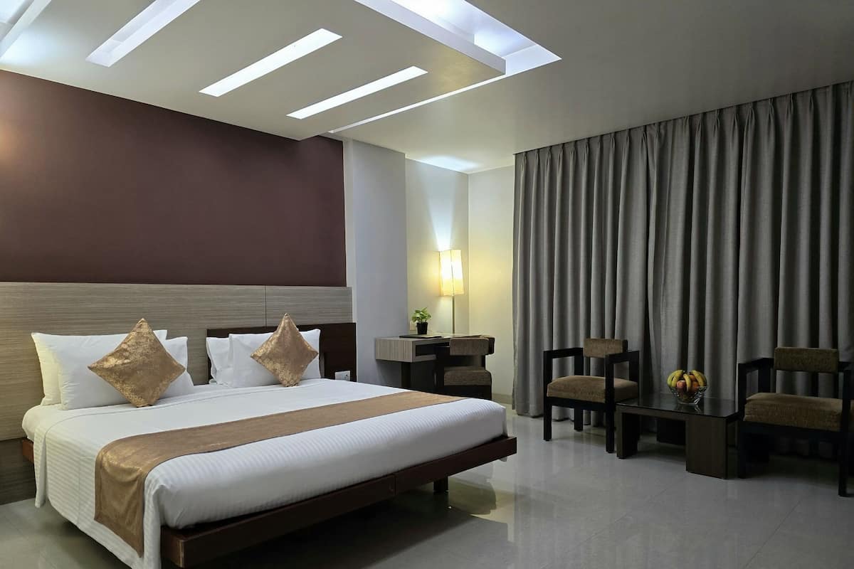 A hotel room with a large bed, a desk, two chairs, a lamp, and a view of the outside through curtains.