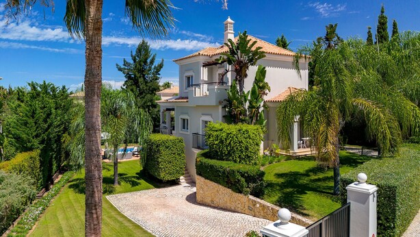 A two-story house with a tiled roof, surrounded by lush greenery and palm trees.