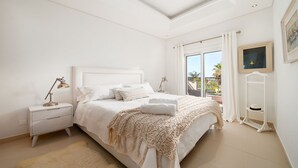 A bedroom with a large bed, white walls, a ceiling with skylights, a nightstand, a lamp, and a view of the outdoors through a sliding glass door.