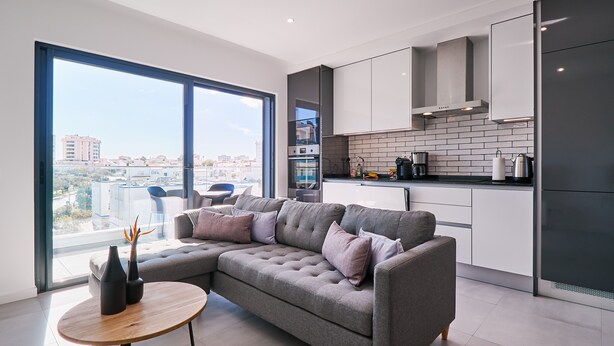 A modern living room with a grey sectional sofa, a round wooden coffee table, and a large window offering a city view. - Terrace Daniel (Portimao)