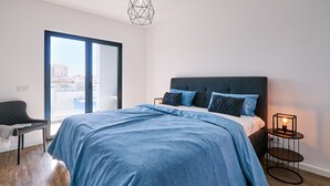 A modern bedroom with a large bed, a chair, a side table with a lamp, and a view of buildings through a large window. - Terrace Daniel (Portimao)