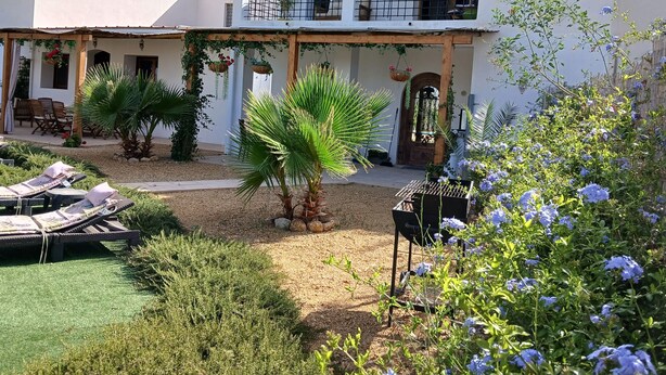Un bâtiment à deux étages doté d’un balcon, d’un espace extérieur aménagé avec des sièges et d’un jardin agrémenté de plantes et de fleurs.