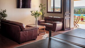 Un salon avec un canapé en cuir marron, une table basse et un téléviseur fixé au mur. Une grande fenêtre offre une vue sur une piscine et des collines.