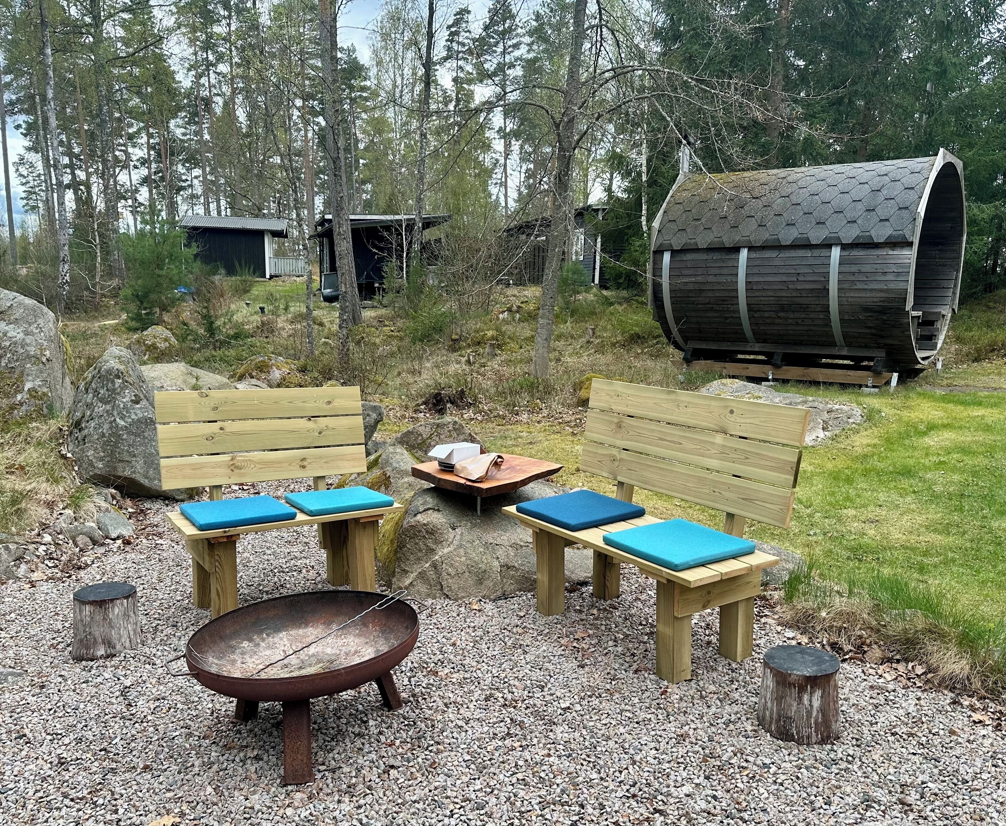 A rustic outdoor seating area with wooden benches and a fire pit, surrounded by trees and a wooden barrel.