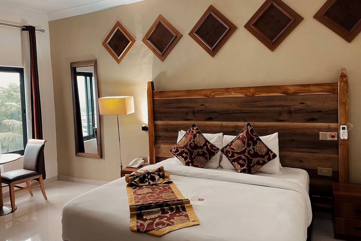 A hotel room with a large bed, a wooden headboard, a bedside table with a lamp, and a view of the outdoors through a window.