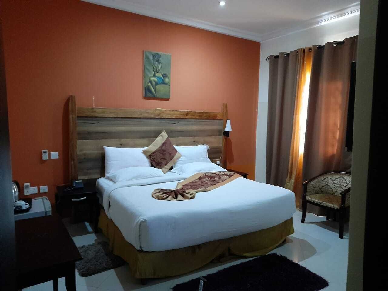 Una habitación de hotel con cabecera de madera, cama con ropa blanca, manta marrón, una silla y un cuadro en la pared.