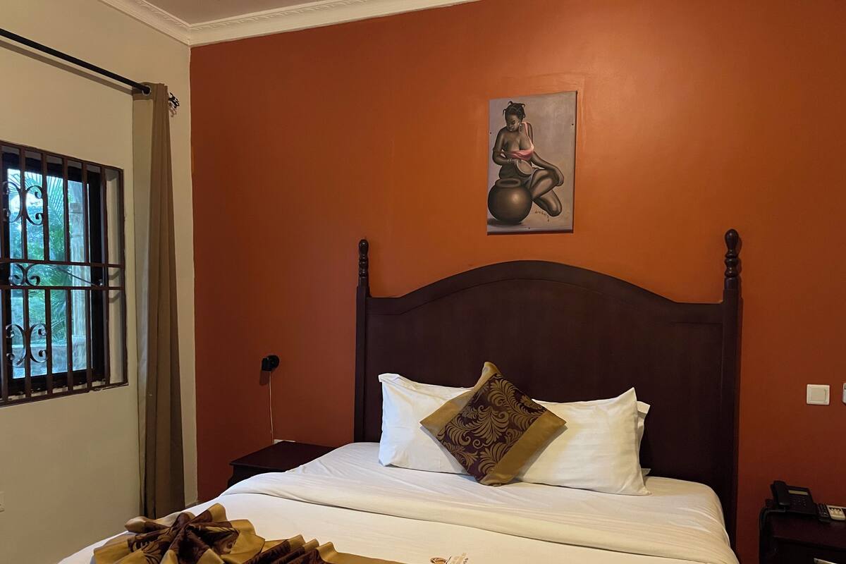 A neatly made bed with white linens and a decorative throw, a wooden headboard, and a painting of a woman on the wall.