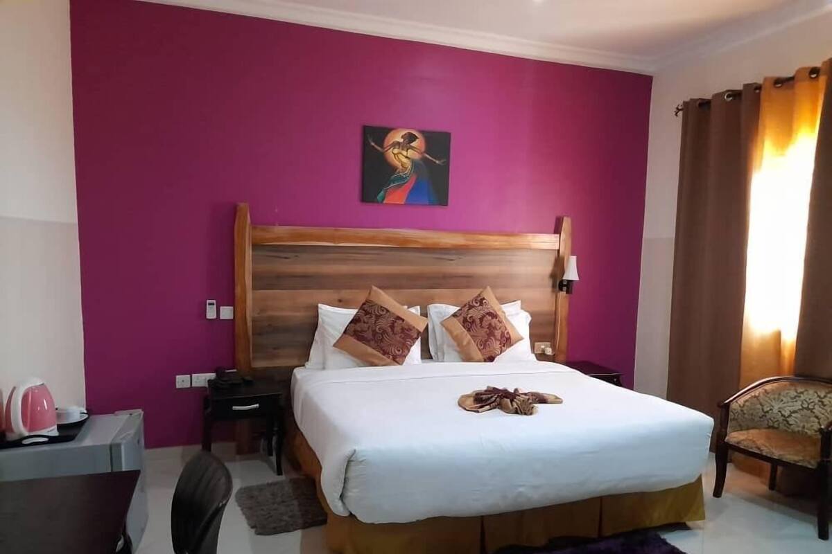 A hotel room with a wooden headboard, a bed with white linens, a purple rug, a chair, and a desk with a pink mug.