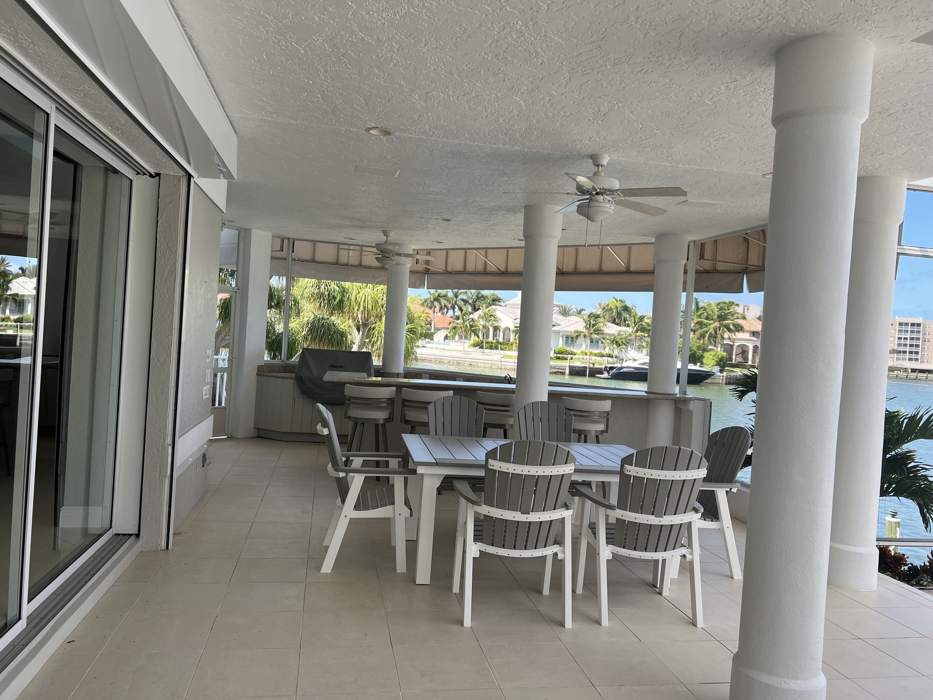 A covered outdoor dining area with white chairs and a table, a ceiling fan, and a view of a body of water and palm trees.