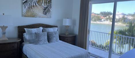 A bedroom with a large bed, two nightstands, a view of a body of water, and a balcony.