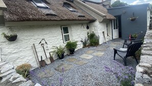 A stone house with a thatched roof, a small garden with gravel paths, and a patio area with a chair.