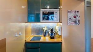 Een compacte keuken met een magnetron, een kleine koelkast en een koffieapparaat. Er hangt een poster aan de muur en op het aanrecht ligt een set bestek.