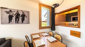 Een eetgedeelte met een houten tafel voor vier personen, een schilderij van skiërs aan de muur, en een keuken die door een open deur zichtbaar is.