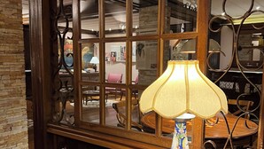 Un bureau en bois avec un livre ouvert, une lampe décorative et une fenêtre donnant sur une pièce où l’on aperçoit des chaises et une table.