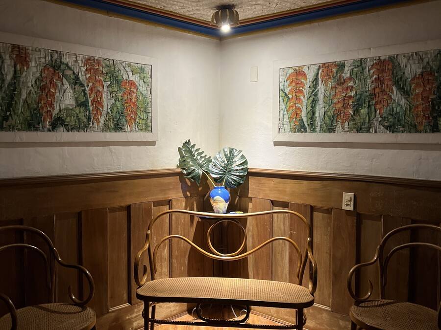 A room with a wooden bench and two chairs, a plant on a shelf, and two paintings on the walls.