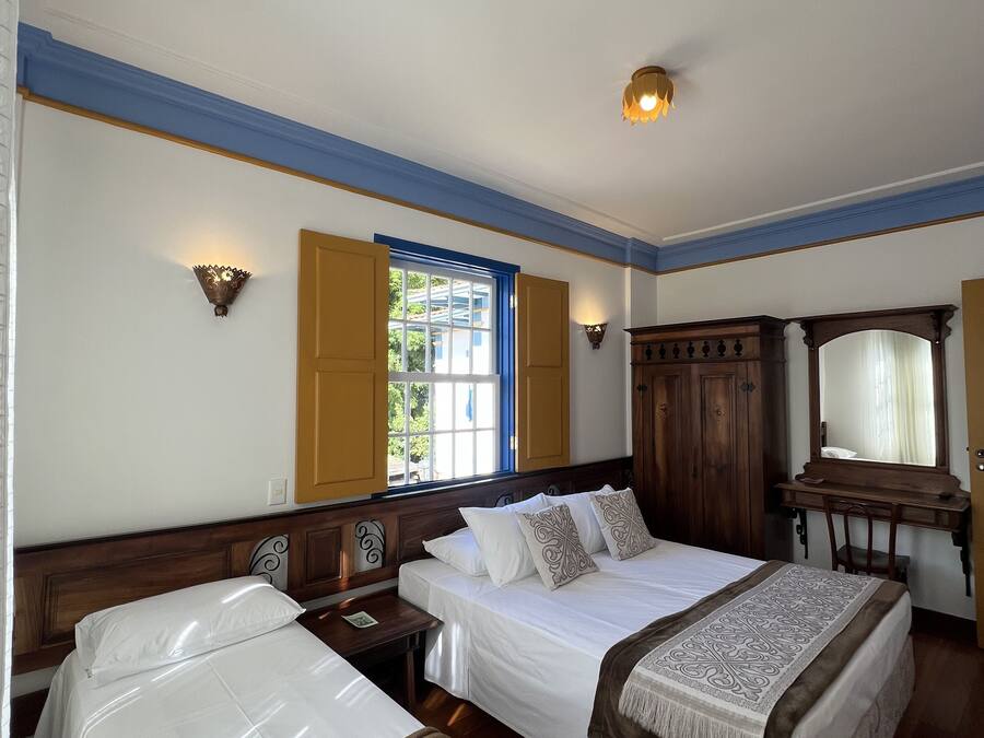 A hotel room with two single beds, a wooden desk, a mirror, and a window with shutters.