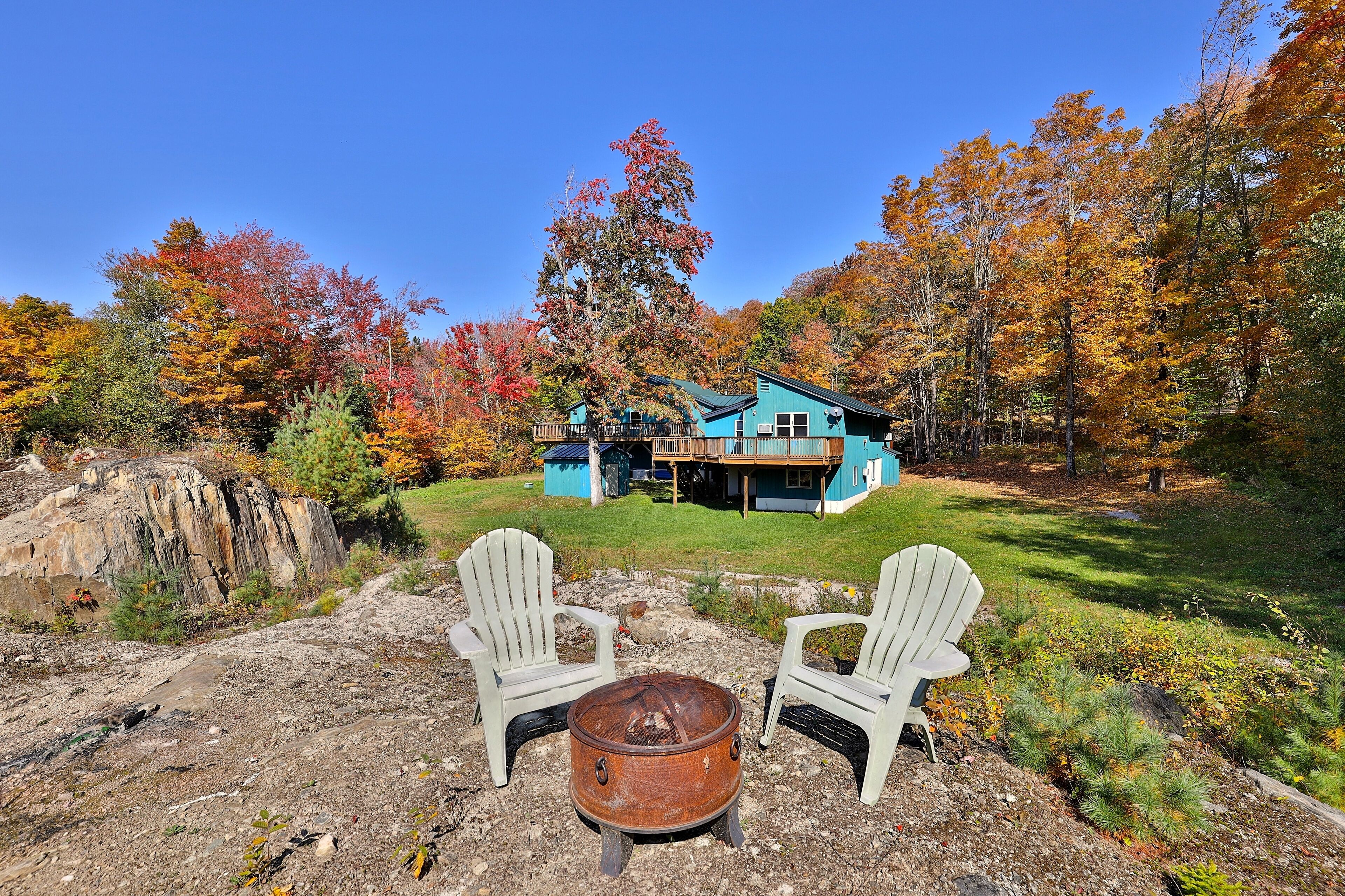 A rustic cabin with a blue exterior, surrounded by autumn foliage. There are two Adirondack chairs and a metal fire pit in the foreground.