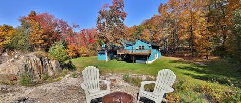 A rustic cabin with a blue exterior, surrounded by autumn foliage. There are two Adirondack chairs and a metal fire pit in the foreground.