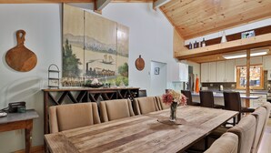 Un comedor amplio con una mesa de madera y bancos acolchados, un cuadro grande en la pared y una cocina al fondo.