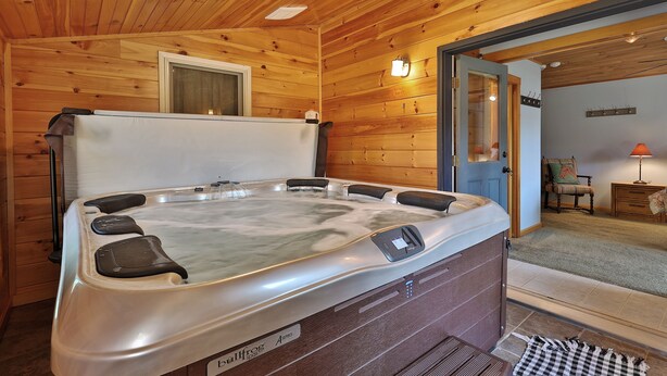 Interior de una cabaña de madera con un jacuzzi, paredes de madera, una ventana y una puerta que da a otra habitación.