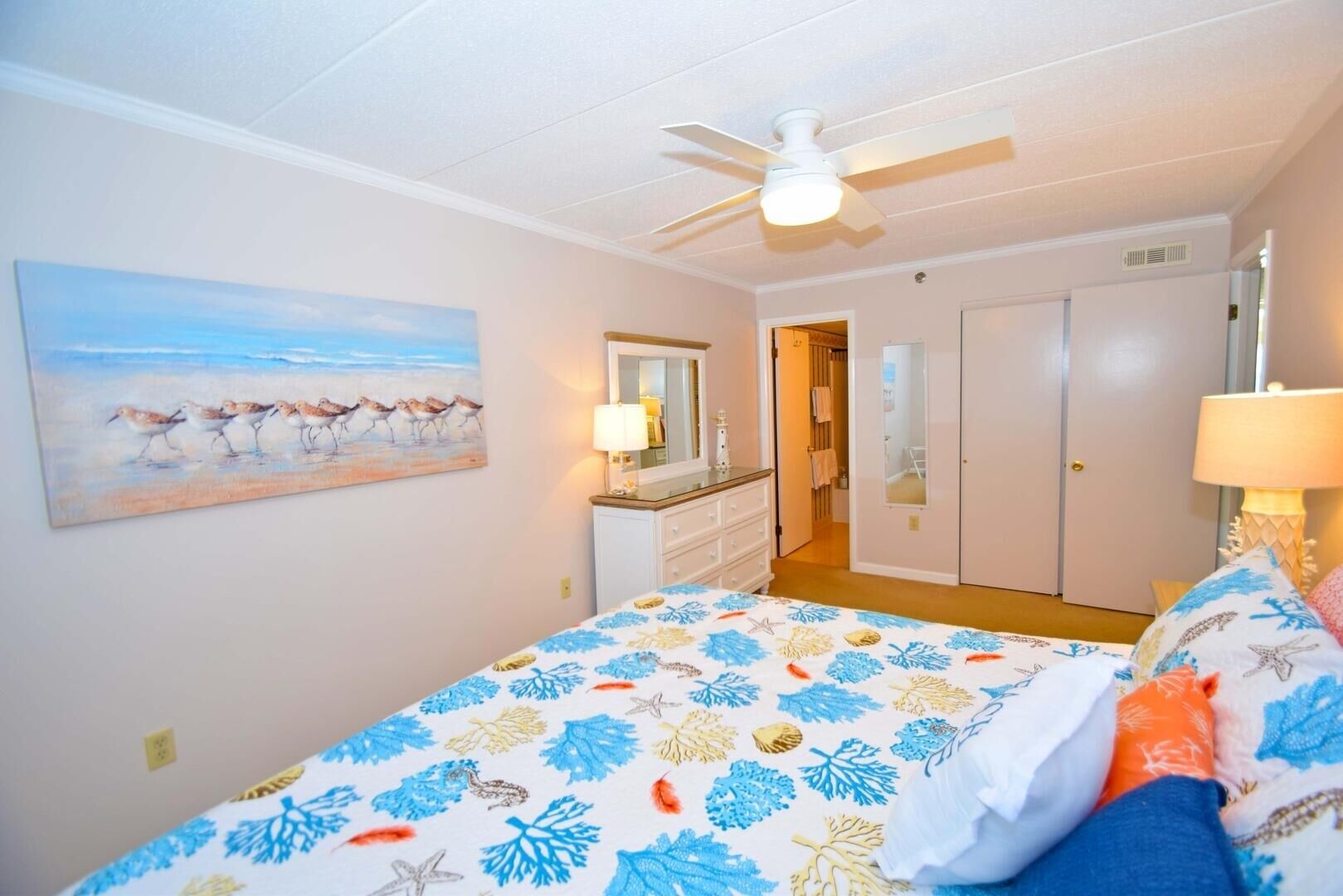 Un dormitorio con cama, ventilador de techo, un cuadro de pájaros en la pared y una puerta que da a otra habitación.
