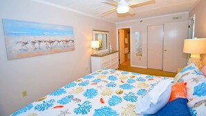 A bedroom with a bed, a ceiling fan, a painting of birds on the wall, and a door leading to another room.