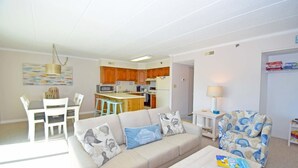 A living room with a sofa, a coffee table, and a patterned armchair. There is a kitchen area with wooden cabinets and a microwave.
