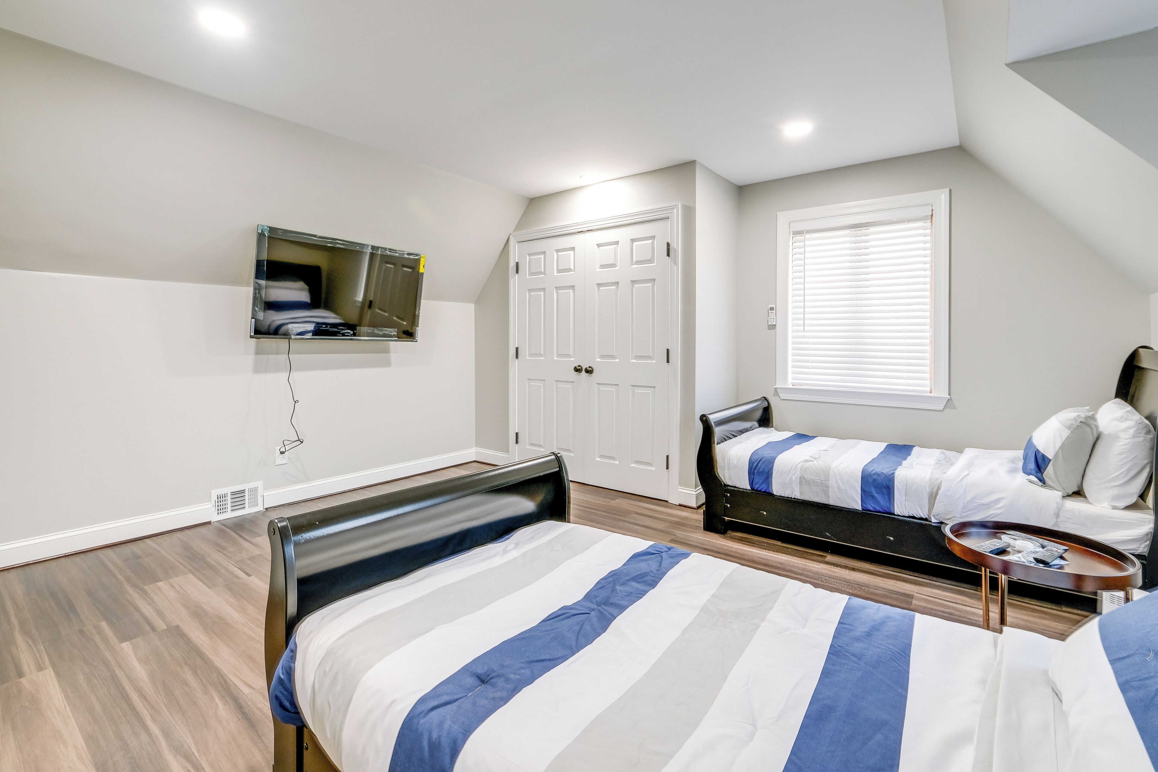 A bedroom with two beds, a TV mounted on the wall, a window with blinds, and a door.