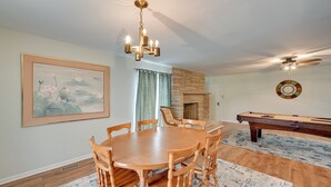 A dining area with a wooden table and chairs, a chandelier, a framed painting, a fireplace, and a pool table.