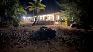 Une propriété située en bord de mer, avec un bateau sur le sable, des palmiers et un bâtiment dont les fenêtres sont illuminées.